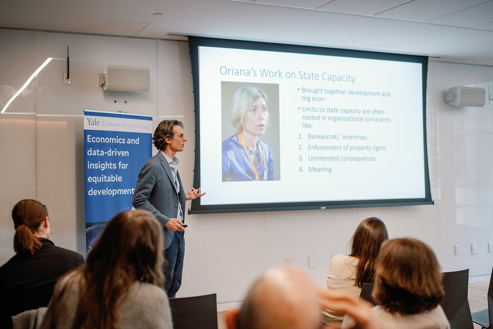A man in a grey suit speaks to an audience with a presentation slide behind him