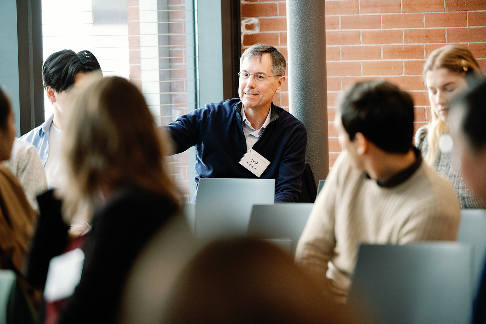 A man in a dark blue sweater asks a question from the back of a classroom
