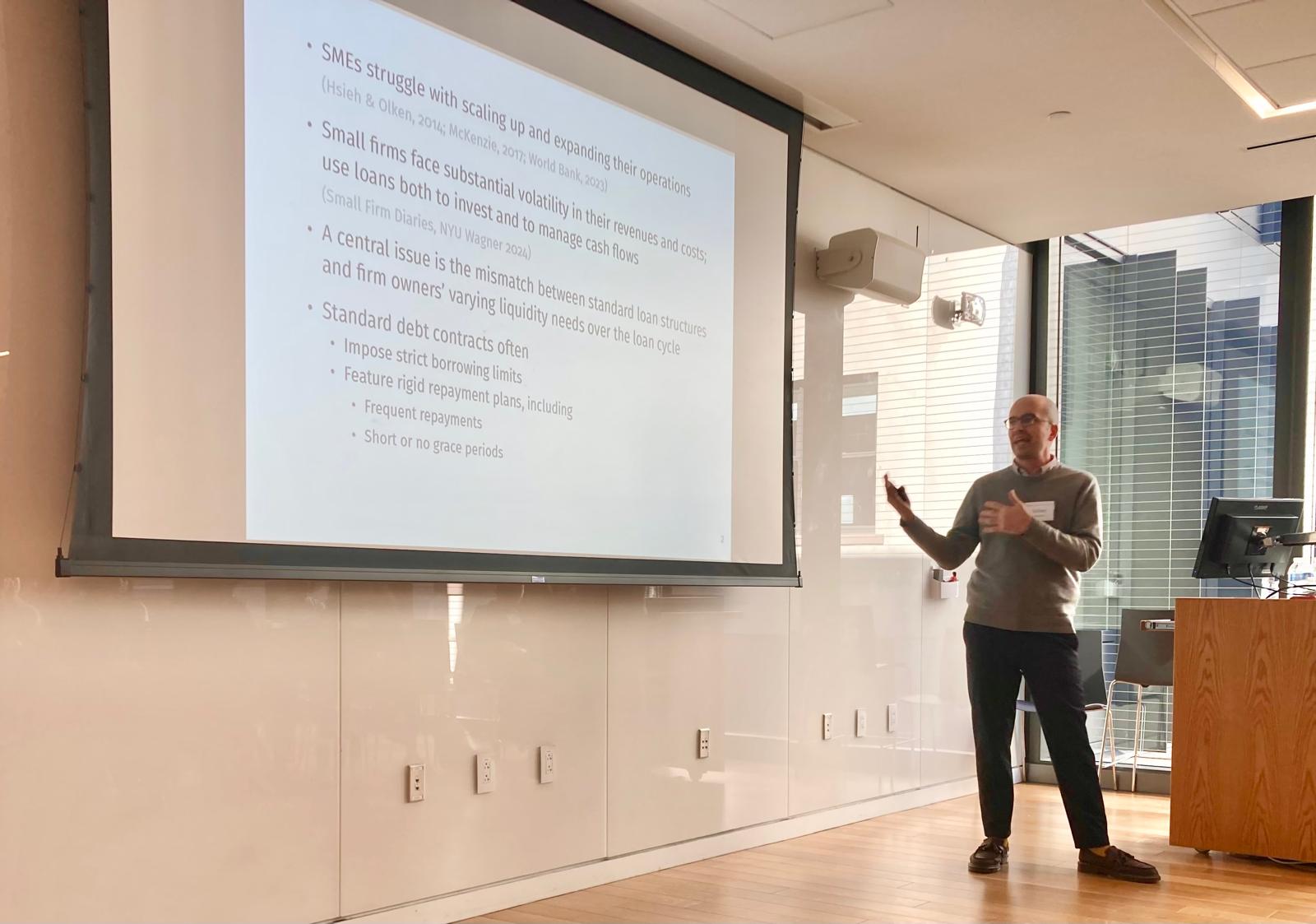 A man in a grey sweater speaks at the front of a classroom