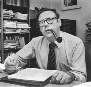 Arthur M. Okun at desk with pipe (1928-1980)
