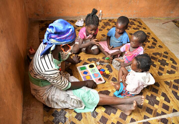Photo of 4 young children sitting in a semi-circle learning shapes with a female adult.